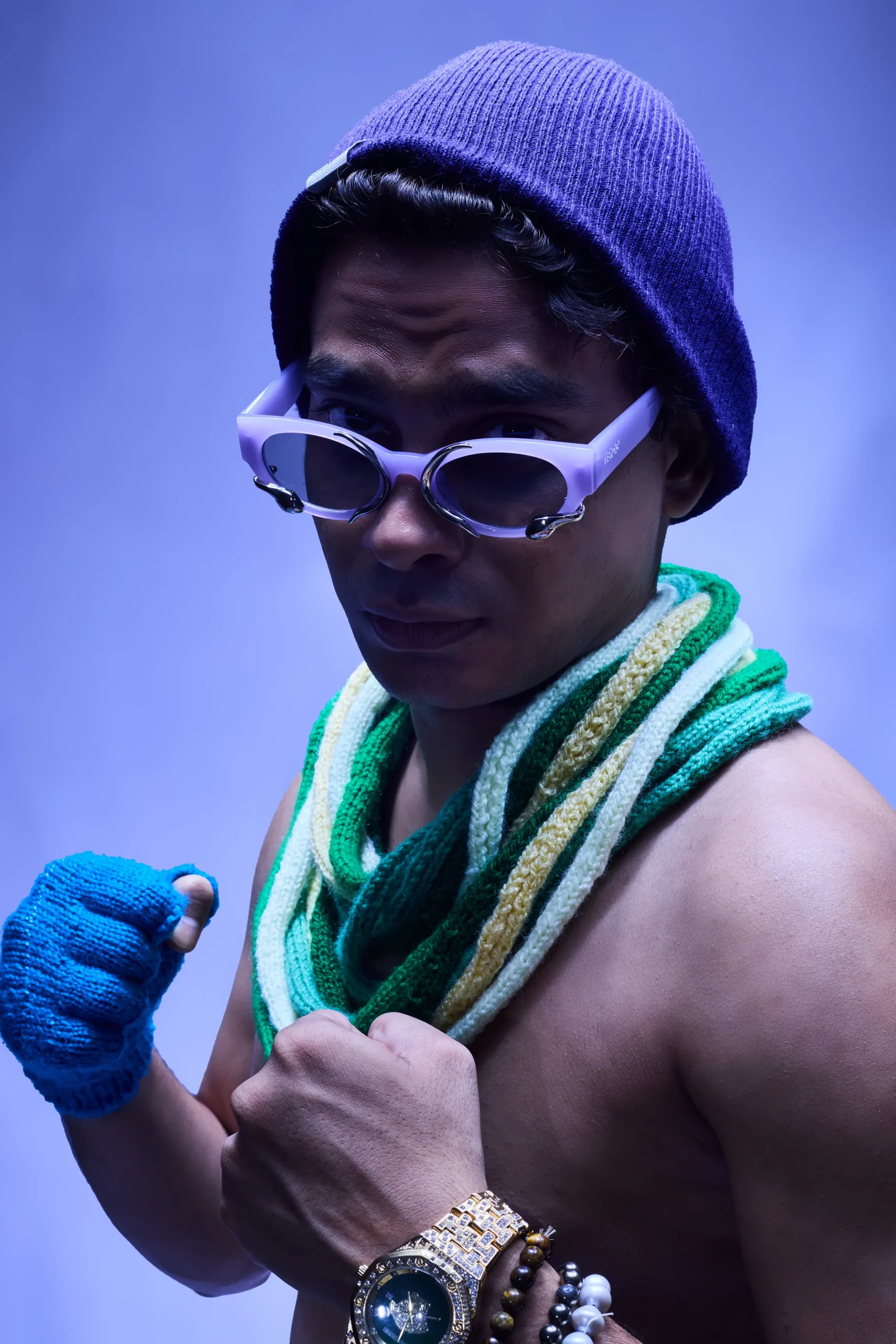 A shirtless person wearing a purple beanie, chunky green and yellow knit scarf, blue fingerless gloves, and multiple bracelets and a large gold watch, poses with fists raised in a playful, confident stance.