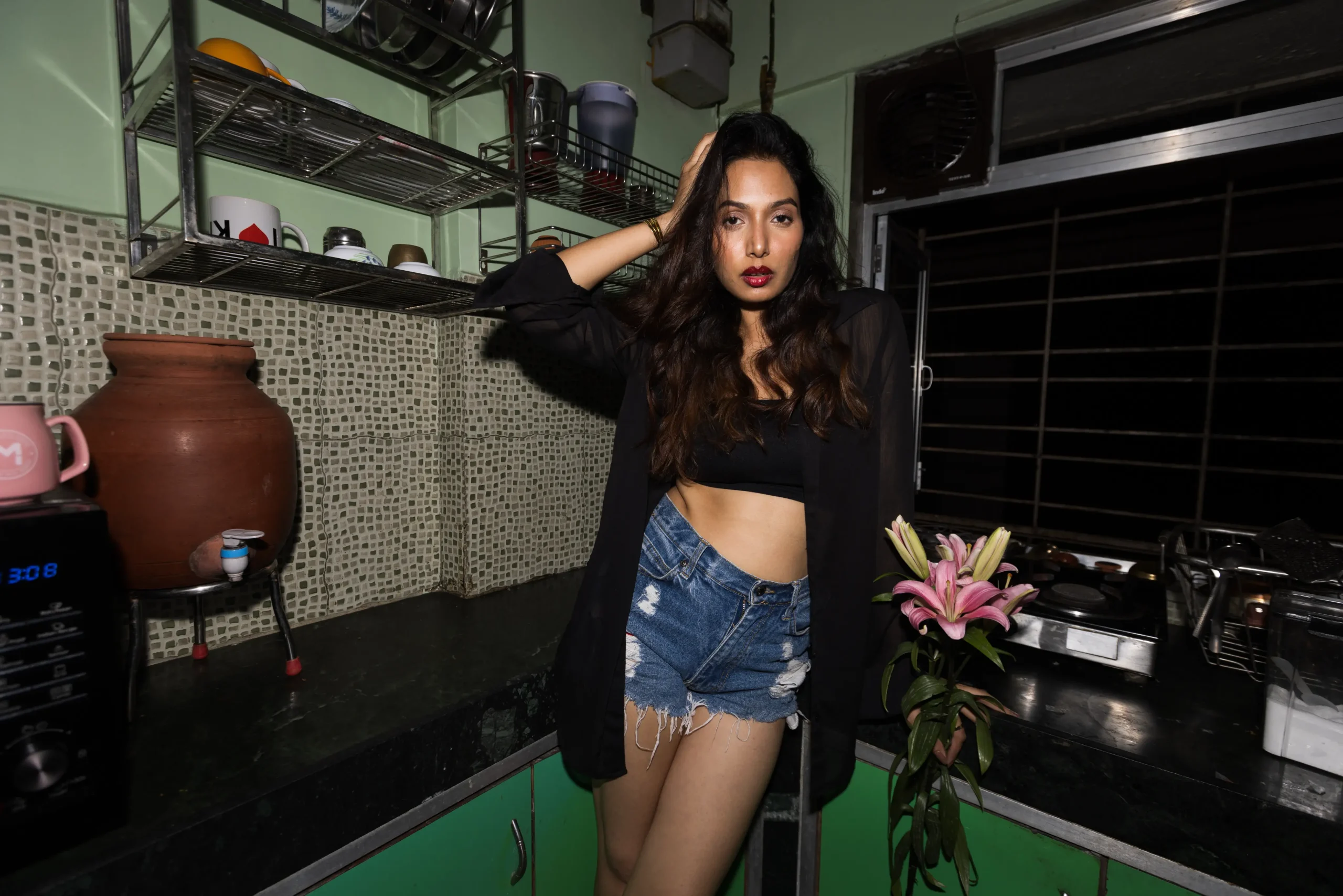 A woman with long wavy hair, wearing a black sheer shirt over a black crop top and high-waisted denim shorts, is sitting on a kitchen counter. The kitchen has green walls, metal racks with utensils, a clay water pot, and a gas stove.