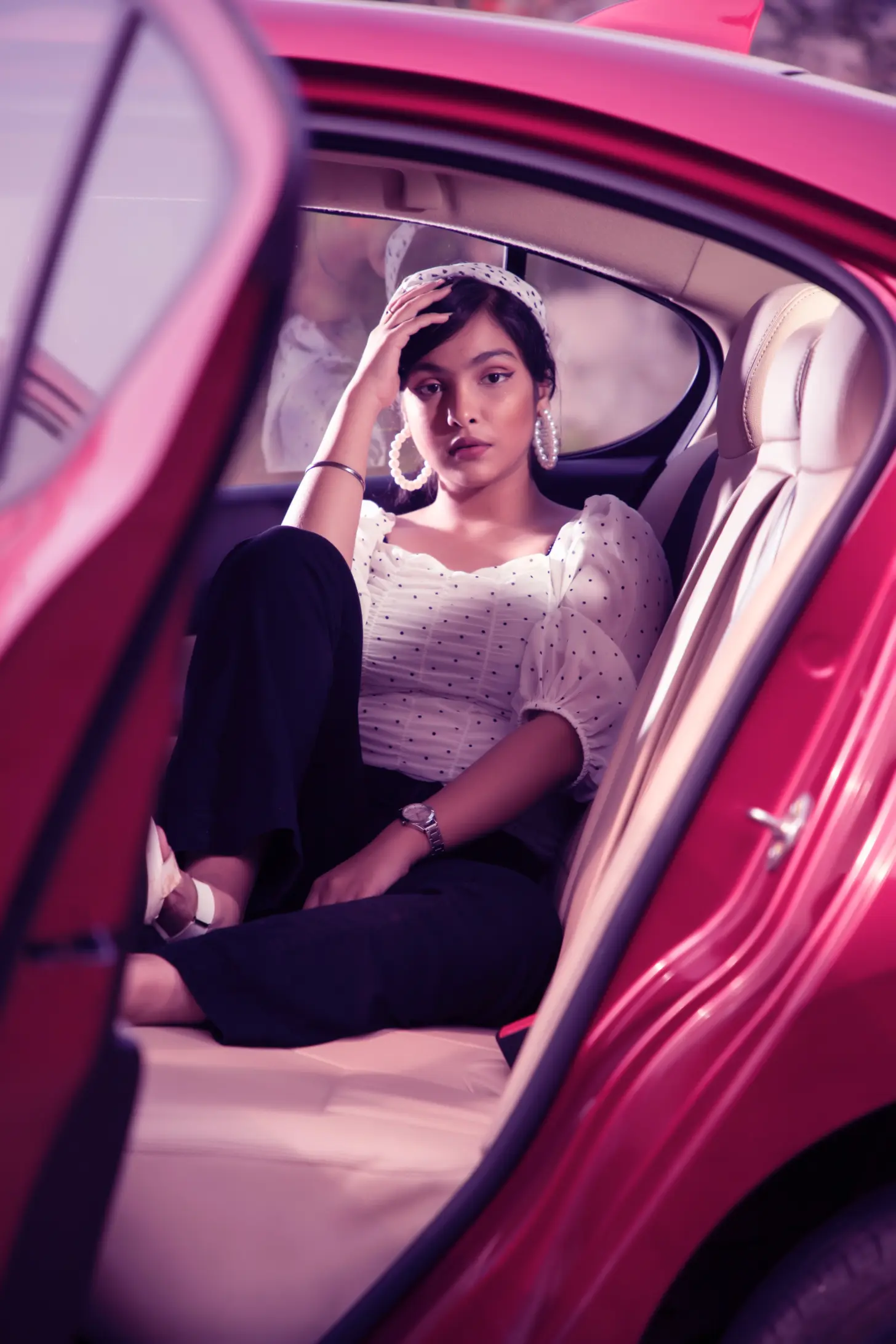 A woman wearing a white polka dot blouse, dark pants, and white sandals sits comfortably in the back seat of a red car with the door open. She accessorizes with a headband, large hoop earrings, a wristwatch, and a bracelet.