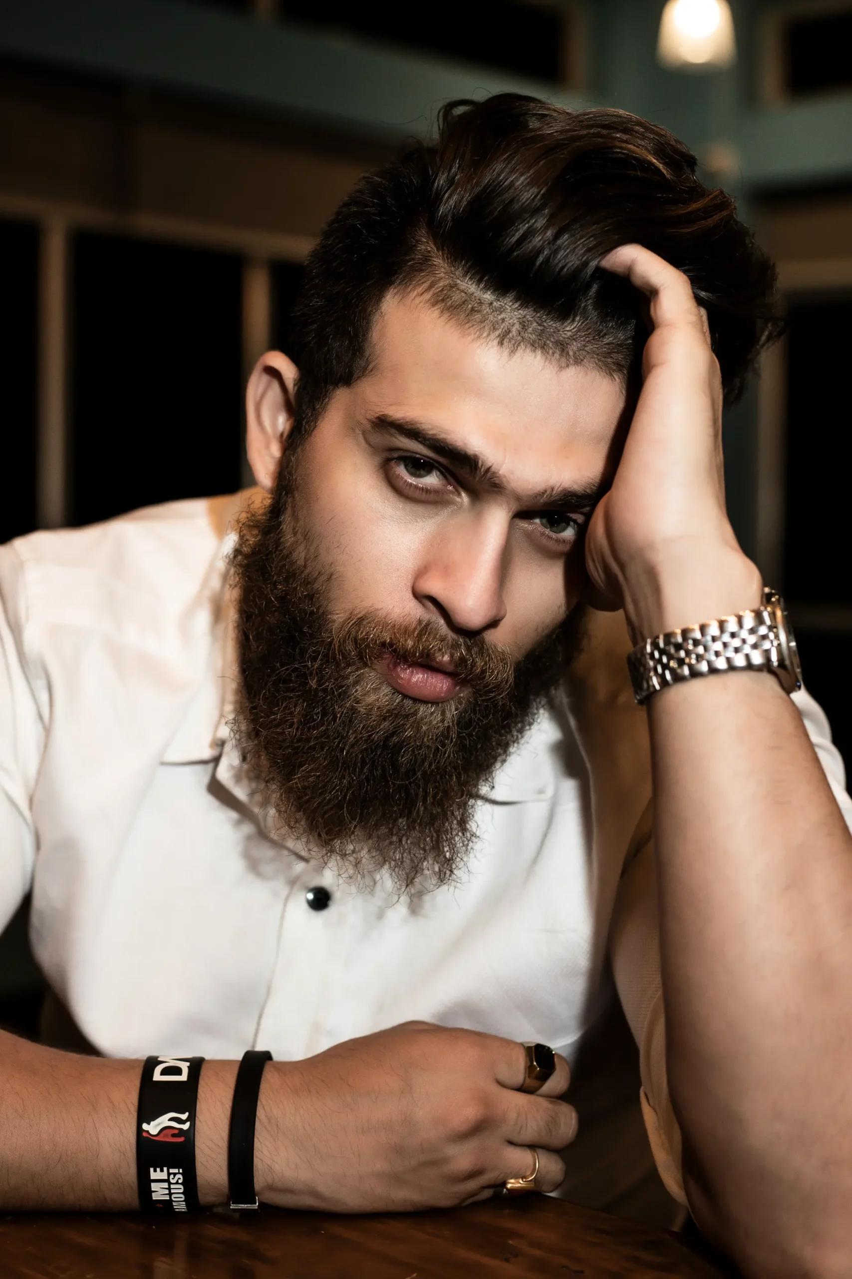 Bearded man in a white shirt leaning on a table, gazing intensely at the camera with his hand in his hair.